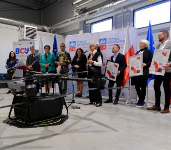 Grupa ośmiu dorosłych osób stoi na podium w dużej hali, uczestnicząc w ceremonii. Z przodu widoczny jest jeden z dużych dronów, a po prawej baner BCJ SPEDYCJA. Osoby trzymają pamiątkowe dyplomy i bukiet kwiatów.