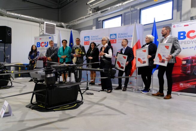 Grupa ośmiu dorosłych osób stoi na podium w dużej hali, uczestnicząc w ceremonii. Z przodu widoczny jest jeden z dużych dronów, a po prawej baner BCJ SPEDYCJA. Osoby trzymają pamiątkowe dyplomy i bukiet kwiatów.