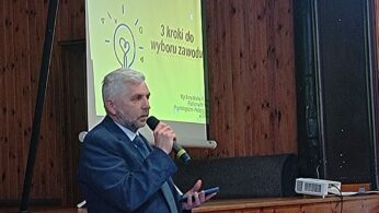 Prelegent ubrany w granatowy garnitur przemawia na tle slajdu wyświetlanego na ekranie. Na slajdzie znajduje się napis „3 kroki do wyboru zawodu” oraz symbol zapalonej żarówki.