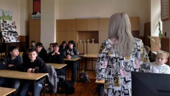 Ujęcie na salę lekcyjną z rzędami ławek oraz krzeseł, przy których siedzą młodzi uczniowie. Naprzeciwko uczniów występuje młoda kobieta. Uczniowie wykazują na twarzach zainteresowanie tematem spotkania.