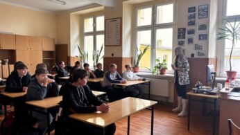 Ujęcie z boku na salę lekcyjną z rzędami ławek oraz krzeseł, przy których siedzą młodzi uczniowie. Naprzeciwko uczniów występuje młoda kobieta, a obok niej rozłożony na ławce zestaw gogli umożliwiających doświadczenie wirtualnej rzeczywistości – gogle VR. Uczniowie wykazują na twarzach zainteresowanie tematem spotkania.