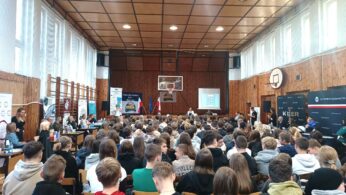 Szkolna sala gimnastyczna wypełniona uczniami klas podstawowych umiejscowionymi w rzędach na krzesłach ustawionych naprzeciwko sceny. Po bokach Sali znajdują się ławki stanowiące miejsce prezentacji firm będących lokalnymi pracodawcami.