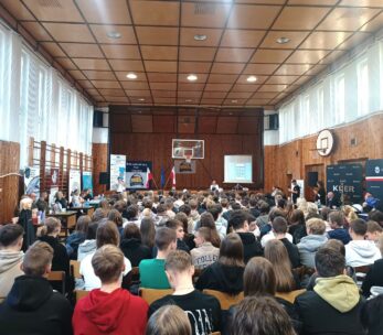 Szkolna sala gimnastyczna wypełniona uczniami klas podstawowych umiejscowionymi w rzędach na krzesłach ustawionych naprzeciwko sceny. Po bokach Sali znajdują się ławki stanowiące miejsce prezentacji firm będących lokalnymi pracodawcami.