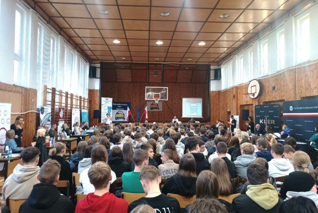 Szkolna sala gimnastyczna wypełniona uczniami klas podstawowych umiejscowionymi w rzędach na krzesłach ustawionych naprzeciwko sceny. Po bokach Sali znajdują się ławki stanowiące miejsce prezentacji firm będących lokalnymi pracodawcami.