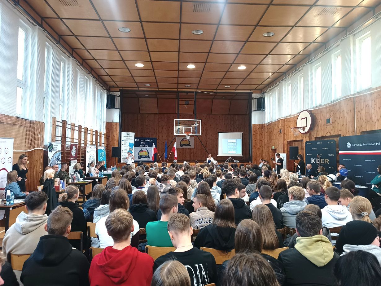 Szkolna sala gimnastyczna wypełniona uczniami klas podstawowych umiejscowionymi w rzędach na krzesłach ustawionych naprzeciwko sceny. Po bokach Sali znajdują się ławki stanowiące miejsce prezentacji firm będących lokalnymi pracodawcami.