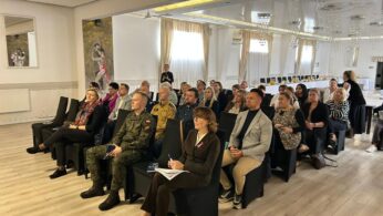 Duża grupa osób siedzi na czarnych krzesłach w rzędach, słuchając prelegenta, który stoi w głębi sali. Sala jest jasna, ma drewnianą podłogę i błyszczący sufit z licznymi lampami. Wśród uczestników znajduje się zarówno młodzież, jak i dorośli, w tym osoba w mundurze.