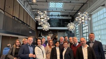 Zdjęcie grupowe kilkunastu osób, kobiet i mężczyzn, zrobione w przestronnym, nowoczesnym biurowcu lub loftowym wnętrzu. W tle widoczny jest wysoki sufit z dużymi przeszkleniami oraz kaskady dużych, srebrnych, lustrzanych lamp. Grupa stoi razem w centrum kadru, uśmiechając się do aparatu, prawdopodobnie po zakończeniu spotkania.