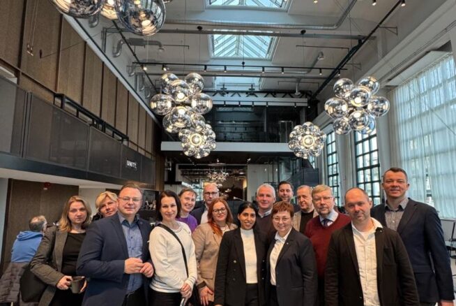 Zdjęcie grupowe kilkunastu osób, kobiet i mężczyzn, zrobione w przestronnym, nowoczesnym biurowcu lub loftowym wnętrzu. W tle widoczny jest wysoki sufit z dużymi przeszkleniami oraz kaskady dużych, srebrnych, lustrzanych lamp. Grupa stoi razem w centrum kadru, uśmiechając się do aparatu, prawdopodobnie po zakończeniu spotkania.