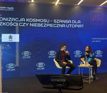 Zdjęcie z panelu dyskusyjnego na dużej scenie z ogromnym ekranem w tle, podczas konferencji "open eyes economy summit". Temat panelu to: "KOLONIZACJA KOSMOSU – SZANSA DLA LUDZKOŚCI CZY NIEBEZPIECZNA UTOPIA?". Dwóch prelegentów, Agata Kołodziejczyk i Jan Brzyk, siedzi na scenie w trakcie debaty.