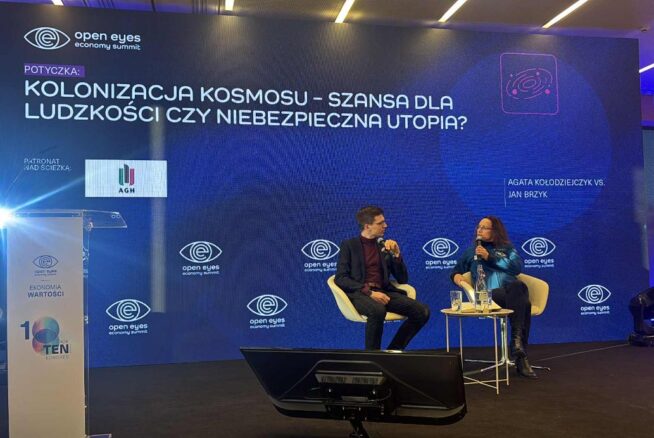 Zdjęcie z panelu dyskusyjnego na dużej scenie z ogromnym ekranem w tle, podczas konferencji "open eyes economy summit". Temat panelu to: "KOLONIZACJA KOSMOSU – SZANSA DLA LUDZKOŚCI CZY NIEBEZPIECZNA UTOPIA?". Dwóch prelegentów, Agata Kołodziejczyk i Jan Brzyk, siedzi na scenie w trakcie debaty.