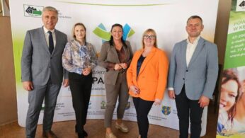 Zdjęcie grupowe pięciu dorosłych osób – trzech mężczyzn i dwie kobiety – stojących na tle bannera informacyjnego. Banner zawiera logo 