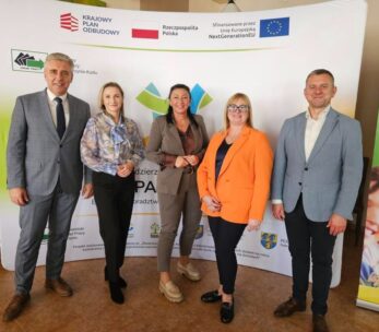 Zdjęcie grupowe pięciu dorosłych osób – trzech mężczyzn i dwie kobiety – stojących na tle bannera informacyjnego. Banner zawiera logo "Krajowego Planu Odbudowy" oraz informacje o finansowaniu z Unii Europejskiej NextGenerationEU. Grupa wygląda na przedstawicieli partnerów i organizatorów spotkania lub projektu.