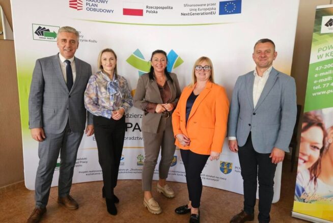 Zdjęcie grupowe pięciu dorosłych osób – trzech mężczyzn i dwie kobiety – stojących na tle bannera informacyjnego. Banner zawiera logo "Krajowego Planu Odbudowy" oraz informacje o finansowaniu z Unii Europejskiej NextGenerationEU. Grupa wygląda na przedstawicieli partnerów i organizatorów spotkania lub projektu.
