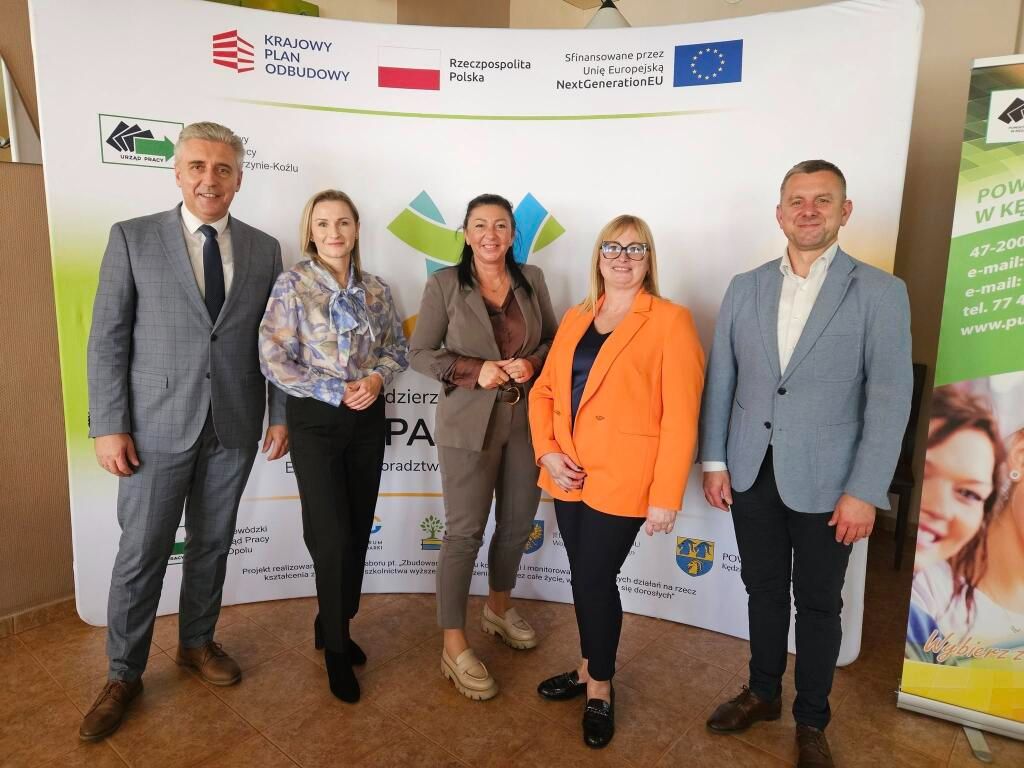 Zdjęcie grupowe pięciu dorosłych osób – trzech mężczyzn i dwie kobiety – stojących na tle bannera informacyjnego. Banner zawiera logo 