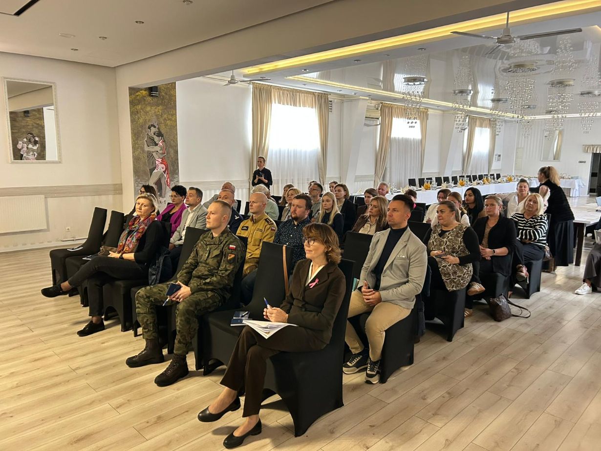 Duża grupa osób siedzi na czarnych krzesłach w rzędach, słuchając prelegenta, który stoi w głębi sali. Sala jest jasna, ma drewnianą podłogę i błyszczący sufit z licznymi lampami. Wśród uczestników znajduje się zarówno młodzież, jak i dorośli, w tym osoba w mundurze.