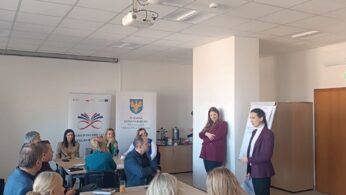 Zdjęcie z warsztatu. Dwie kobiety w bordowych żakietach stoją, prezentując lub prowadząc dyskusję z grupą siedzących przy stołach uczestników. Kobieta po prawej wskazuje na notatki. Uczestnicy siedzą wokół dwóch połączonych stołów, na których znajdują się napoje i materiały.