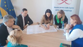 Kilka osób siedzi przy stole, koncentrując się na dużym arkuszu papieru rozłożonym na środku. Jedna z kobiet (w beżowym żakiecie) pisze na arkuszu. Wokół stołu siedzą dwie kobiety w zielonych elementach ubioru oraz dwóch mężczyzn w garniturach. W tle widoczne są flagi, w tym ukraińska, oraz baner.