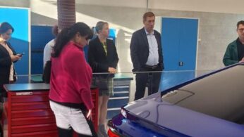 Grupa osób, w tym kobieta w różowej bluzie na pierwszym planie, ogląda tył niebieskiego samochodu Toyota w nowoczesnym warsztacie lub laboratorium. Samochód jest zabezpieczony pomarańczowym pasem. W tle widoczny jest mężczyzna w garniturze i kobieta w żółto-czarnej kratce.