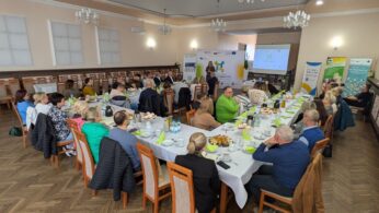 Szerokie ujęcie z góry na spotkanie biznesowe lub szkoleniowe odbywające się w przestronnej sali restauracyjnej lub konferencyjnej. Uczestnicy siedzą przy długim stole ułożonym w kształt litery 