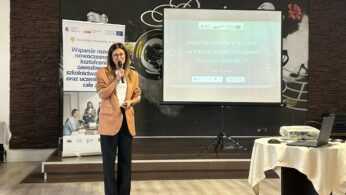 Kobieta w brązowej marynarce i czarnych spodniach stoi przed ekranem projekcyjnym, trzymając mikrofon. Na ekranie widoczny jest tytuł prezentacji w języku polskim: 