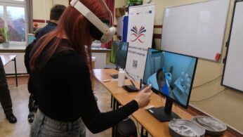 Dziewczyna z rudymi włosami, ubrana na czarno, stoi przy biurku i korzysta z gogli VR oraz kontrolerów. Na monitorze obok wyświetlany jest widok z symulacji wirtualnej, prawdopodobnie związanej z higieną lub medycyną, z dłonią w niebieskiej rękawiczce. W tle widoczny jest rollup promocyjny.