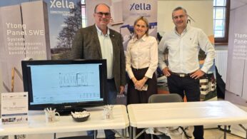 Trzy osoby — dwóch mężczyzn i kobieta — stoją za stołem wystawowym firmy Xella Construction Services, zajmującej się systemami budowlanymi Ytong. Przed nimi na ekranie monitora widoczny jest rysunek techniczny. Na stole stoją kubki i miseczka.