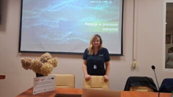Kobieta stojąca za biurkiem w sali konferencyjnej, z lekko pochylonym tułowiem, uśmiechnięta i patrząca w obiektyw. W tle widać duży ekran, na którym wyświetlone jest logo firmy Umicore i hasło 