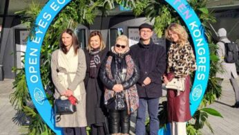 Zdjęcie grupowe pięciu elegancko ubranych osób stojących na zewnątrz przed dużym, przeszklonym budynkiem. Osoby pozują w dużej, okrągłej ramie dekoracyjnej, która jest ozdobiona zielonymi liśćmi i zawiera napis 