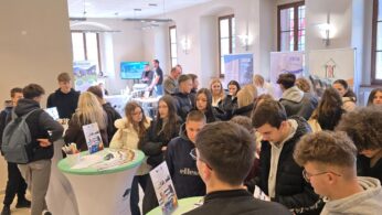 Duże, jasne pomieszczenie wypełnione młodymi ludźmi, którzy stoją przy różnych stoiskach promocyjnych. W centralnej części kadru widać kilka osób rozmawiających przy okrągłych, wysokich stolikach. W tle widoczne są banery reklamowe firm, w tym Xella i TBC Development.