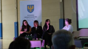 Cztery kobiety siedzą na panelu dyskusyjnym. Jedna z nich trzyma mikrofon i mówi. W tle widać rollup z herbem i nazwiskiem 