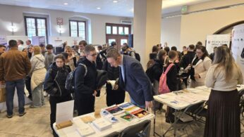 Panoramiczne ujęcie dużej sali, na której odbywają się targi pracy lub edukacji. Wiele osób, głównie młodzieży, gromadzi się przy stoiskach, rozmawiając z ich przedstawicielami. W centrum kadru widać dorosłego mężczyznę pochylającego się nad materiałami promocyjnymi.