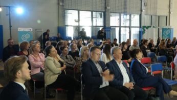 Zdjęcie przedstawiające uczestników konferencji siedzących w rzędach krzeseł na sali z dużymi oknami w tle, przez które wpada światło. Ludzie w różnym wieku i strojach uważnie słuchają prelegenta (niewidocznego). Na pierwszym planie po lewej siedzi mężczyzna w garniturze.