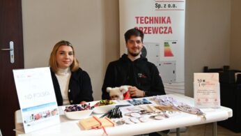 Kobieta i mężczyzna siedzą za stoiskiem firmy ND Polska, specjalizującej się w technice grzewczej. Na stole leży duża ilość gadżetów reklamowych, w tym przypinki, długopisy i słodycze. Mężczyzna trzyma na kolanach małego, białego psa.
