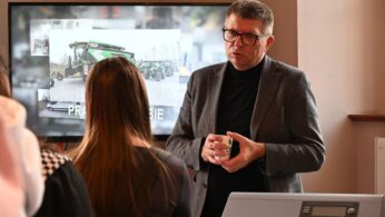 Mężczyzna w okularach i szarej marynarce wygłasza prezentację przed małą grupą słuchaczy. Trzyma w dłoniach mały biały kubek. Za nim widoczny jest duży ekran telewizora, na którym wyświetlany jest obraz maszyn rolniczych, prawdopodobnie firmy Agro-Eko.