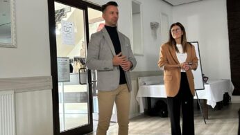 W jasnym pomieszczeniu stoją dwie osoby: mężczyzna w jasnoszarej marynarce i beżowych chinosach oraz kobieta w brązowej marynarce i czarnych spodniach. Stoją w pobliżu drzwi prowadzących na zewnątrz oraz białej tablicy na sztaludze. Tło zdobią lustra w ozdobnych ramach.