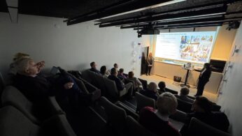 Ujęcie na salę konferencyjną z rzędami siedzeń, przypominającą małą salę kinową. Publiczność, złożona z kilkunastu osób, siedzi w półmroku, obserwując duży ekran projekcyjny. Na ekranie widoczny jest slajd z tekstem i zdjęciami, prawdopodobnie związany z rozwojem kompetencji. Prezentację prowadzą dwie osoby, stojące po prawej stronie ekranu.