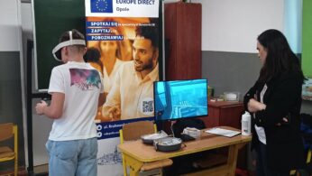 Chłopak w białej koszulce i dżinsach stoi tyłem, używając gogli VR i trzymając kontrolery. Z boku stoi kobieta, obserwując go z założonymi rękami. Na monitorze przed nimi widoczny jest widok z wirtualnej rzeczywistości. Obok wisi baner z logo Europe Direct Opole.