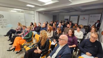 Zdjęcie przedstawia widownię siedzącą na pomarańczowych krzesłach w nowoczesnej sali konferencyjnej, prawdopodobnie podczas szkolenia. Wszyscy uczestnicy są skupieni na prezentacji, która odbywa się poza kadrem. W tle widoczne są plakaty i logo, w tym 