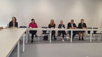 Siedem osób (sześć kobiet i jeden mężczyzna) siedzi za długim, białym stołem w jasnej sali konferencyjnej. Uczestnicy są ubrani w stroje biznesowe i casualowe. W tle widoczny jest rollup z herbem i nazwiskiem 