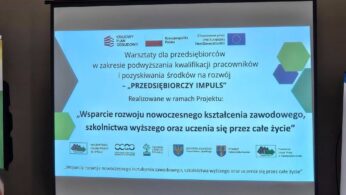 Zdjęcie dużego ekranu projekcyjnego wyświetlającego slajd informacyjny w języku polskim. Tytuł na ekranie to: 