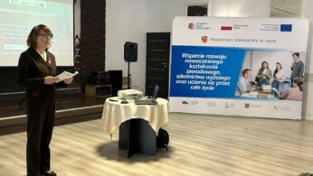 Kobieta w ciemnym kostiumie stoi przed banerem z informacjami o projekcie i logotypami, w tym 