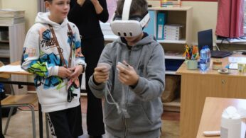 Dwóch chłopców stoi w klasie; jeden z nich ma na głowie gogle VR i obsługuje kontrolery, a drugi stoi obok, patrząc. Chłopiec w goglach jest ubrany w szarą bluzę z kapturem. W tle widoczne są pomoce naukowe i biurko.