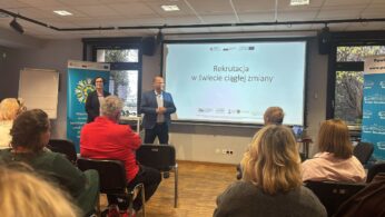 Mężczyzna w jasnoniebieskiej marynarce przemawia do publiczności stojąc obok kobiety, przed ekranem z napisem 