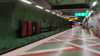 Szerokie ujęcie peronu metra z bardzo charakterystycznym, artystycznym wystrojem. Ściany są malowane na ciemną zieleń i wyglądają jak jaskinia lub surowe skały, z czerwonymi, rzeźbionymi akcentami dekoracyjnymi. Podłoga peronu ma wzór czerwono-białych pasów na zielonym tle, a w oddali widać zbliżający się pociąg. Jest to najprawdopodobniej jedna ze słynnych stacji sztokholmskiego metra.