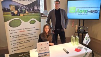 Mężczyzna i młoda kobieta siedzą lub stoją za stoiskiem firmy Agro-Eko, dystrybutora maszyn rolniczych. Za nimi znajduje się duży baner reklamowy z wizerunkiem ciągnika. Obok nich stoi włączony telewizor prezentujący logo firmy.