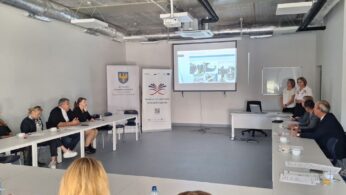 Spotkanie w jasnej sali konferencyjnej. Dwie kobiety stoją przy rzutniku i prowadzą prezentację dla grupy siedzących uczestników. Na slajdzie widoczne są zdjęcia z różnymi scenami z pracy. W tle widać rollupy promujące Województwo Opolskie i inicjatywy 