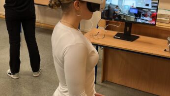 Dziewczyna w białej bluzce z długim rękawem i szarych spodniach dresowych stoi, używając gogli VR i kontrolerów. Na monitorze obok wyświetla się zbliżenie na dłoń w niebieskiej rękawiczce, obsługującą zawór w wirtualnej przestrzeni. W tle widać szkolną tablicę z listą zawodów i imion.