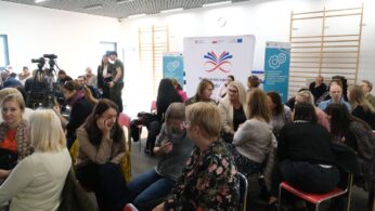 Zdjęcie z sali konferencyjnej, przedstawiające grupę uczestników siedzących i rozmawiających w trakcie przerwy lub networkingu. W tle widoczne są osoby obsługujące sprzęt filmowy. W centrum znajduje się baner z logo i hasłem 