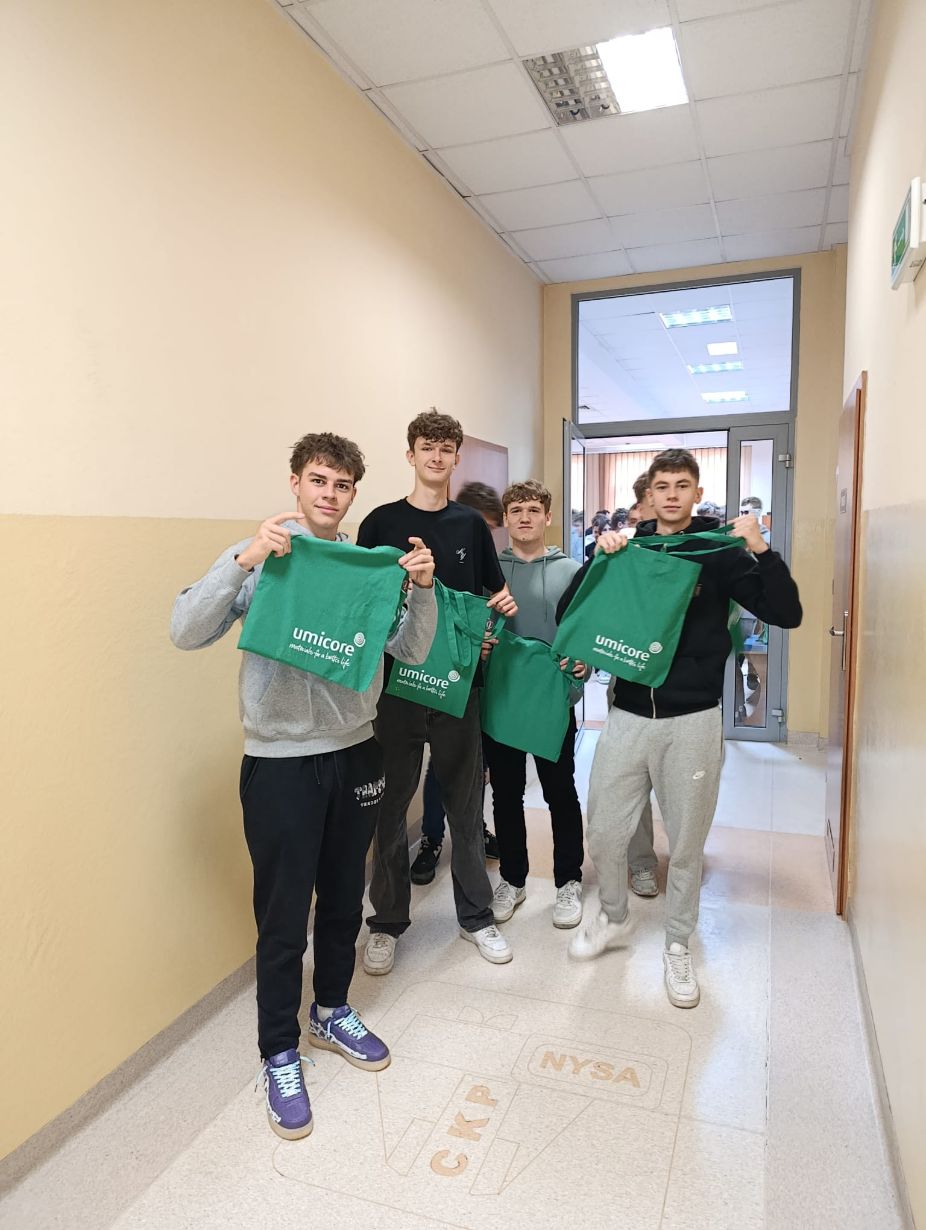 Czterech młodych mężczyzn stoi w korytarzu, uśmiechając się do aparatu i trzymając zielone torby z logo firmy Umicore. W tle widać otwarte drzwi do pomieszczenia. Na podłodze widoczny jest napis 