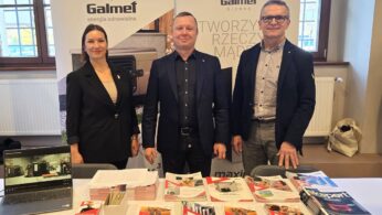 Dwóch mężczyzn i kobieta stoją za stołem wystawowym firmy Galmet, producenta urządzeń grzewczych. Na stole leży duża ilość broszur, ulotek oraz widać otwarty laptop. W tle widoczne są banery reklamowe firmy.